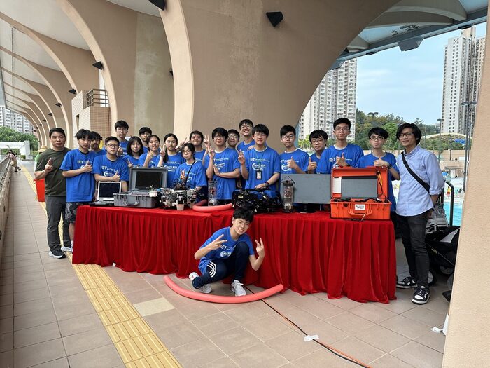 The team behind their equipment tables at a poolside stand, in IET and MATE ROV team shirts